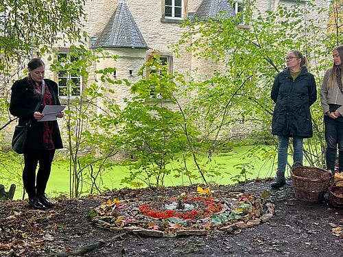 Drei Kolleginnen aus dem Hospiz Hagen stehen am Wasserschloss Werdringen. In der Mitte ist ein Mandala aus Naturmatrialien